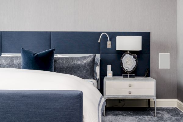 A modern bedroom features a bed with a blue headboard and white linens, a nightstand with a lamp, and decorative elements including pillows and decor.
