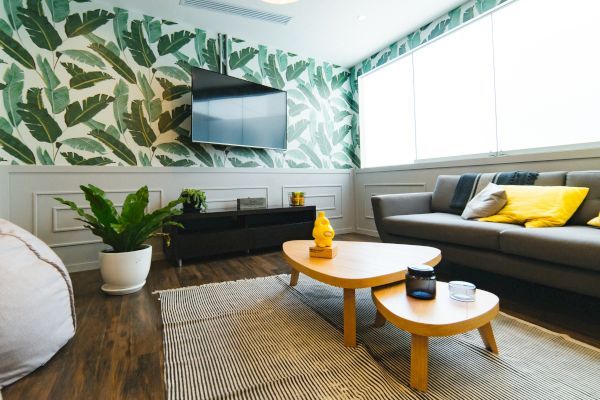 A modern living room with a green leafy wallpaper, gray couch, yellow cushions, wooden coffee table, plant, and wall-mounted TV near a large window.