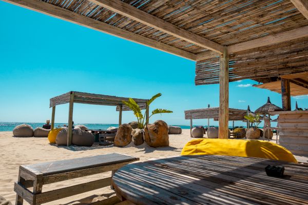 A beachside setting features wooden cabanas, outdoor seating with bean bags, and a clear blue sky. The sand and ocean are visible in the background.