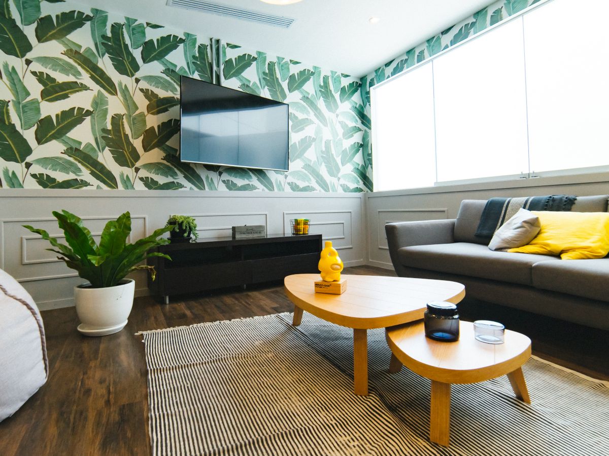 A modern living room with tropical wallpaper, a gray sofa with yellow pillows, a wooden coffee table, large windows, and indoor plants.