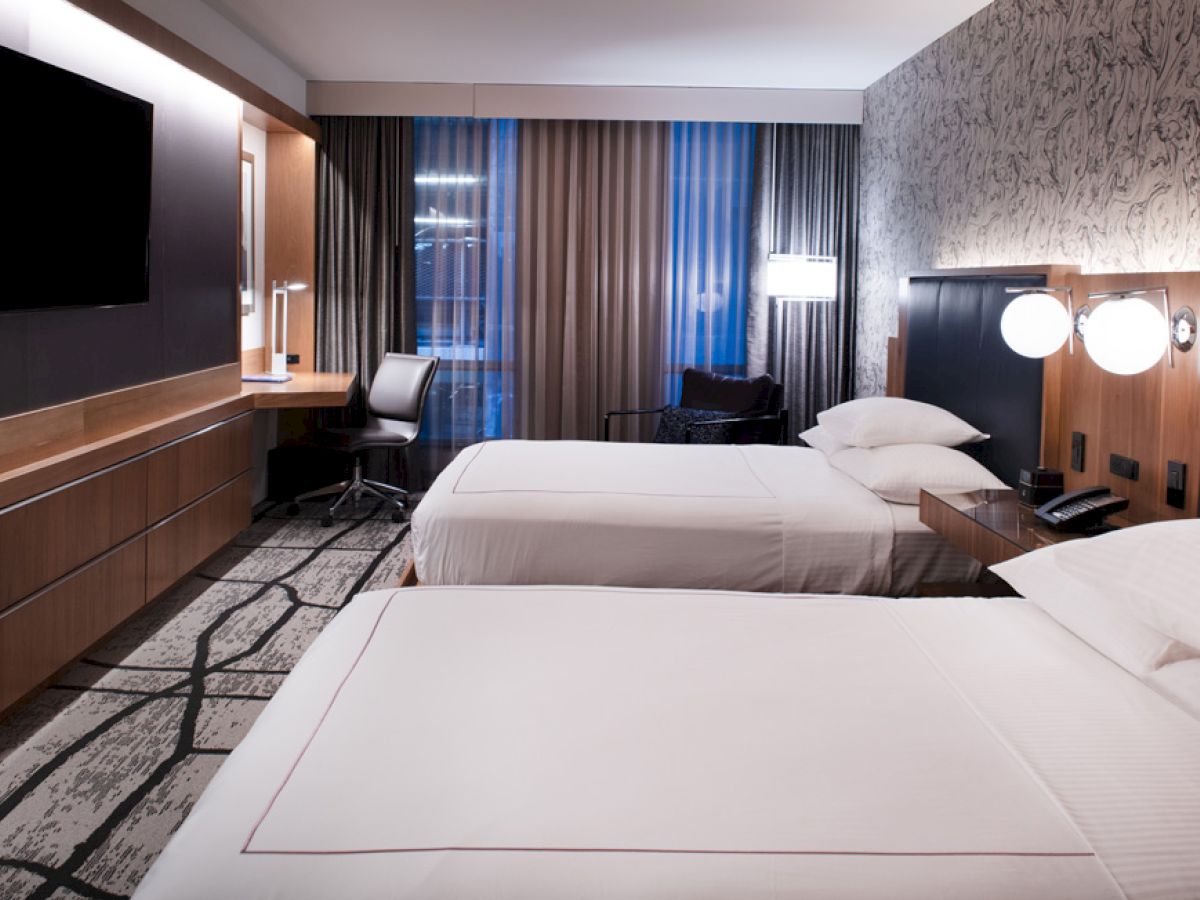 The image shows a modern hotel room with two beds, a TV, a desk, and lamps. The room has stylish decor and large windows with curtains.