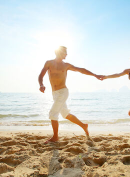 A man and woman are holding hands and running on the beach, with the ocean and sun in the background.