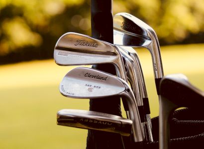 The image shows a set of golf clubs, including brands like Titleist and Cleveland, standing in a golf bag outdoors on a golf course.