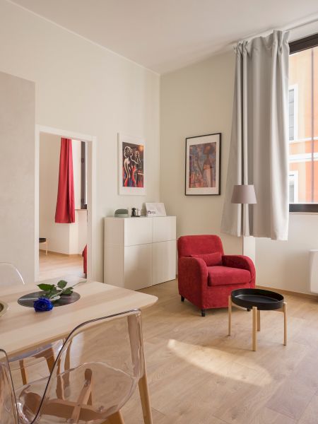 A modern living room with a red chair, a checkered sofa, a dining table, and kitchen appliances. The room is well-lit by a large window.