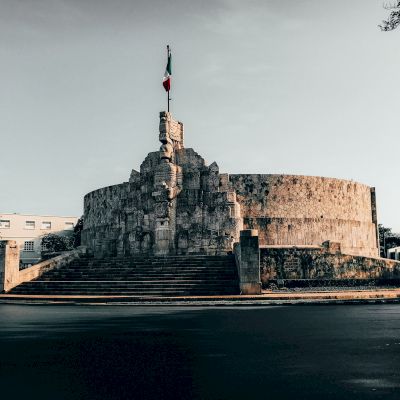 The image shows a large, circular stone monument with a staircase, adorned with sculptures and topped with a flag in an urban setting.