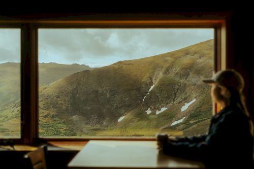 A person sits at a table looking out a window at a mountainous landscape, with patches of snow visible on the slopes and clouds in the sky.