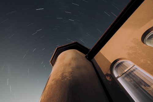 A long exposure photo of a building at night, capturing circular star trails in the sky, with portions of windows visible.