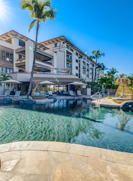 A resort with a large pool, sun loungers, palm trees, and a multi-story building in the background under a bright blue sky.