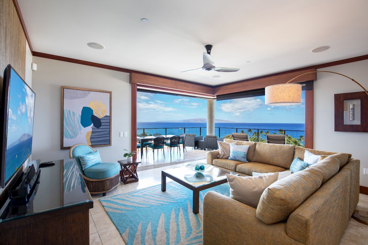 A cozy living room with beige furniture, a TV, modern decor, and a stunning ocean view through large windows and a balcony.