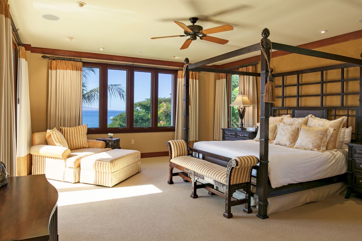 A luxurious bedroom with a four-poster bed, cushioned armchair, large windows, ceiling fan, and ocean view outside.