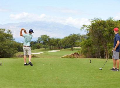 Two men playing golf on a lush, green course with mountains in the background. One swings a club while the other watches.