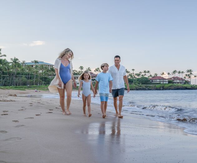 A family of four walks along a sunny beach, holding hands and wading in shallow water near the shoreline.