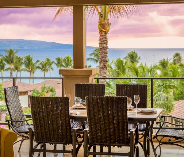 A patio dining set overlooks a serene ocean view with palm trees and mountains in the background under a colorful sky during sunset.