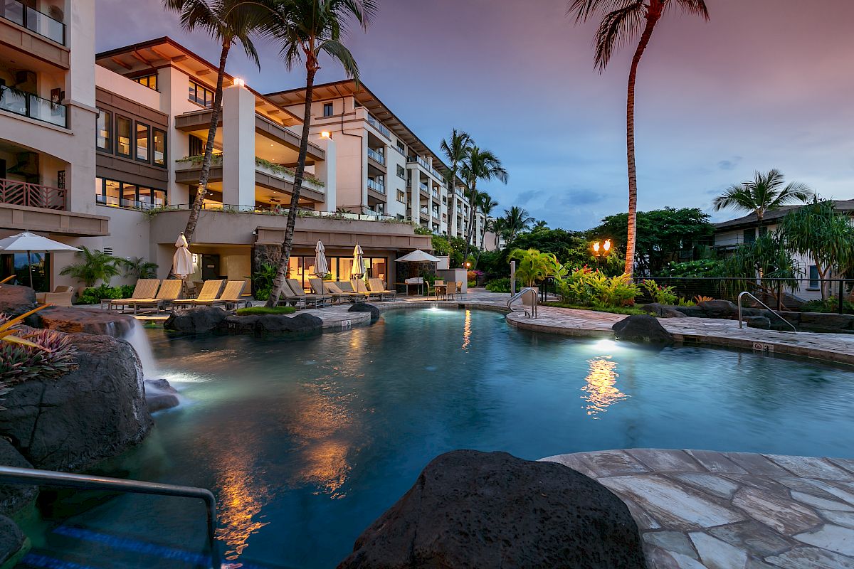 A luxurious resort with a swimming pool, surrounded by palm trees and modern buildings, during the evening with a serene atmosphere.