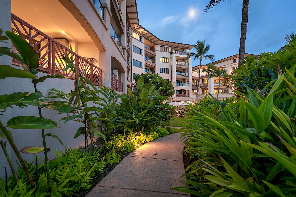 A path lined with lush plants leads to a multi-story building with a tropical setting, captured during evening with lights illuminating the walkway.