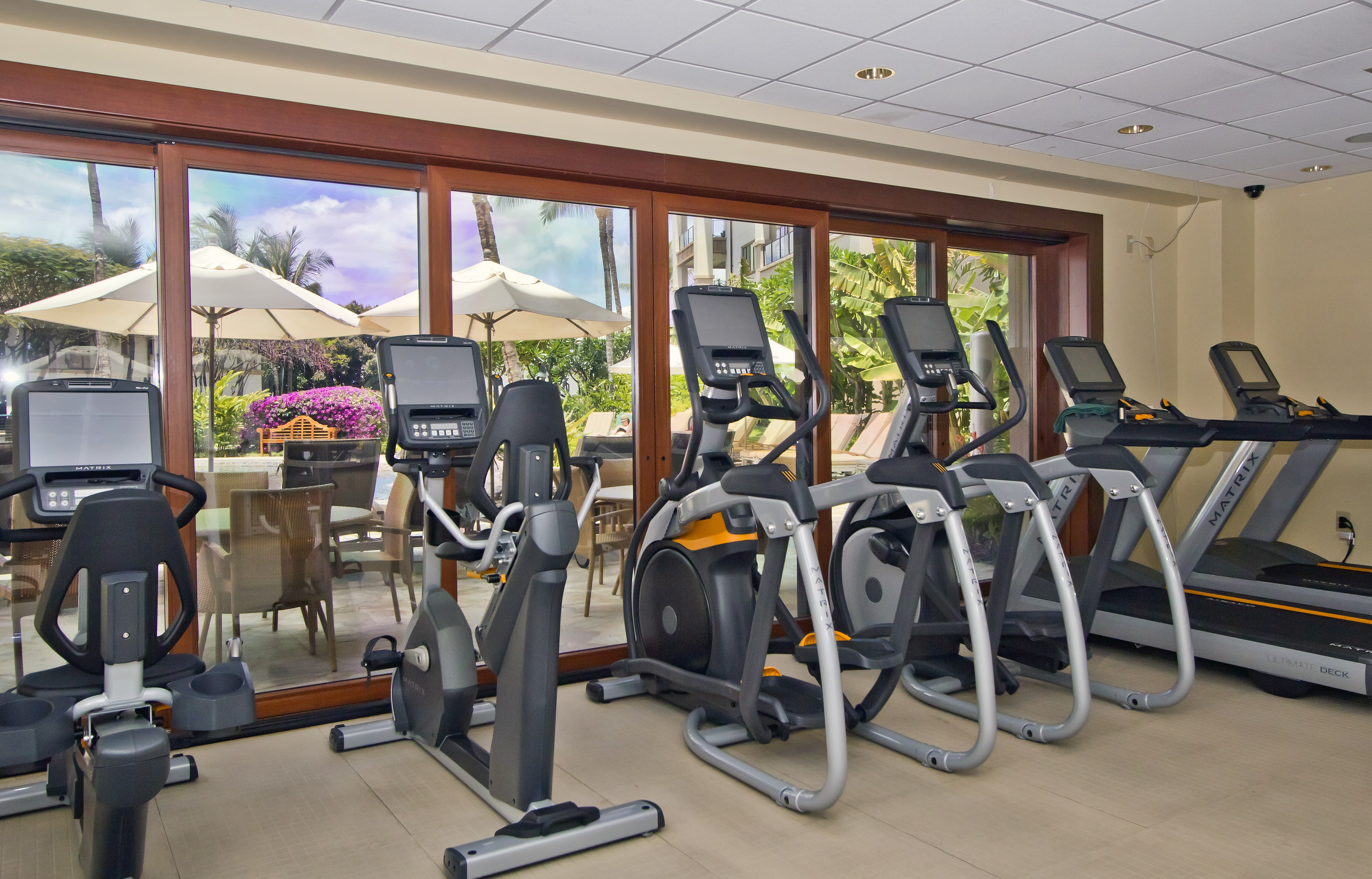 The image shows a gym with various exercise machines like stationary bikes and treadmills, next to glass doors overlooking an outdoor patio.