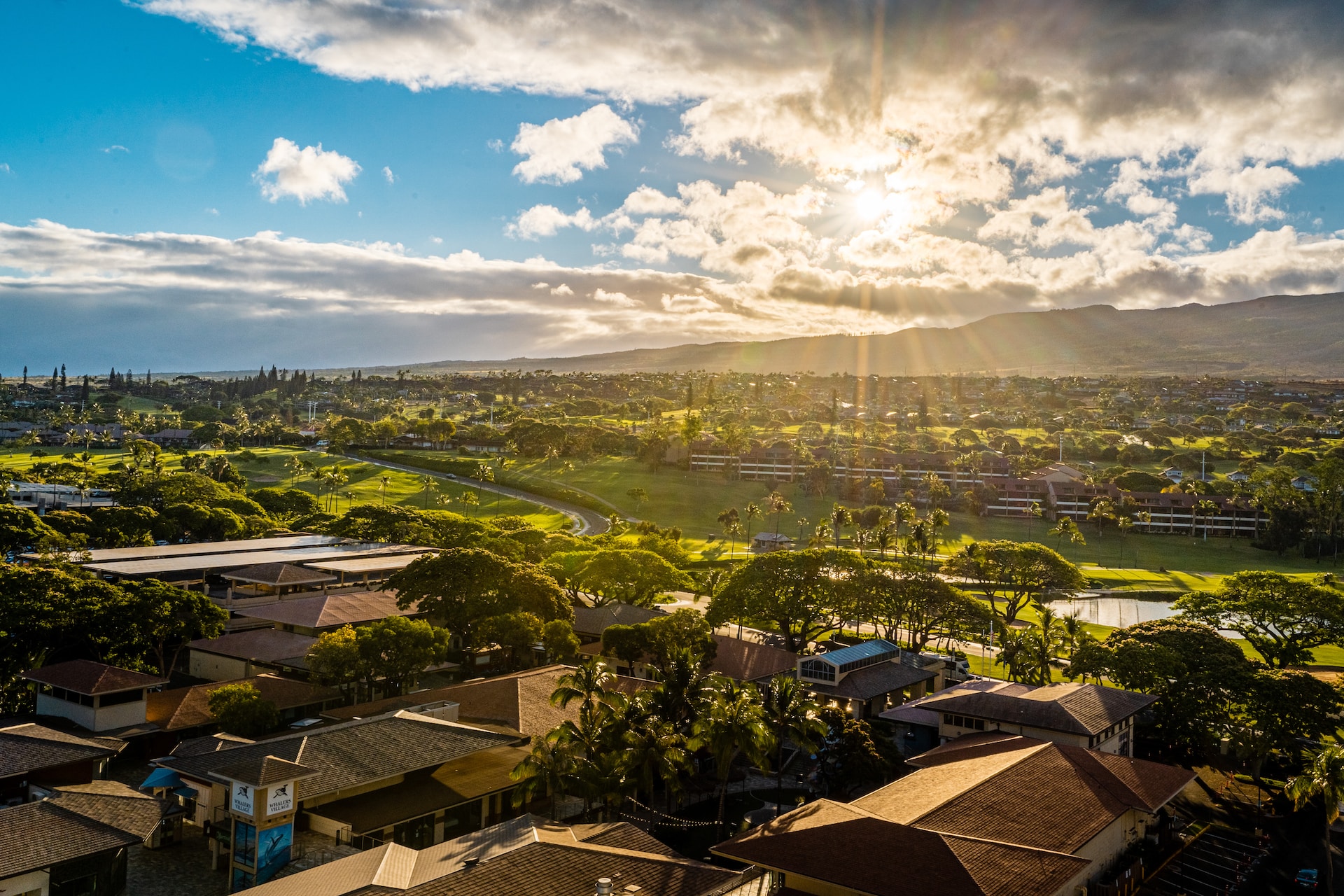 The image shows a scenic landscape of a town with houses, lush greenery, and mountains in the background under a partly cloudy sky with sunlight.