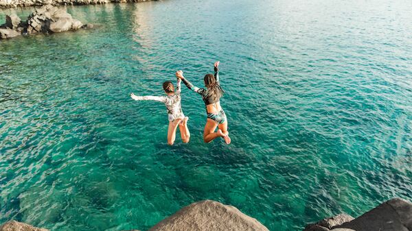 Two people jumping off rocks into clear blue water, holding hands, with a scenic rocky shoreline in the background.