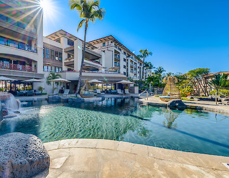 Hotel resort pool area with sunlit palm trees, modern buildings, lounge chairs, and clear blue water at a sunny day.
