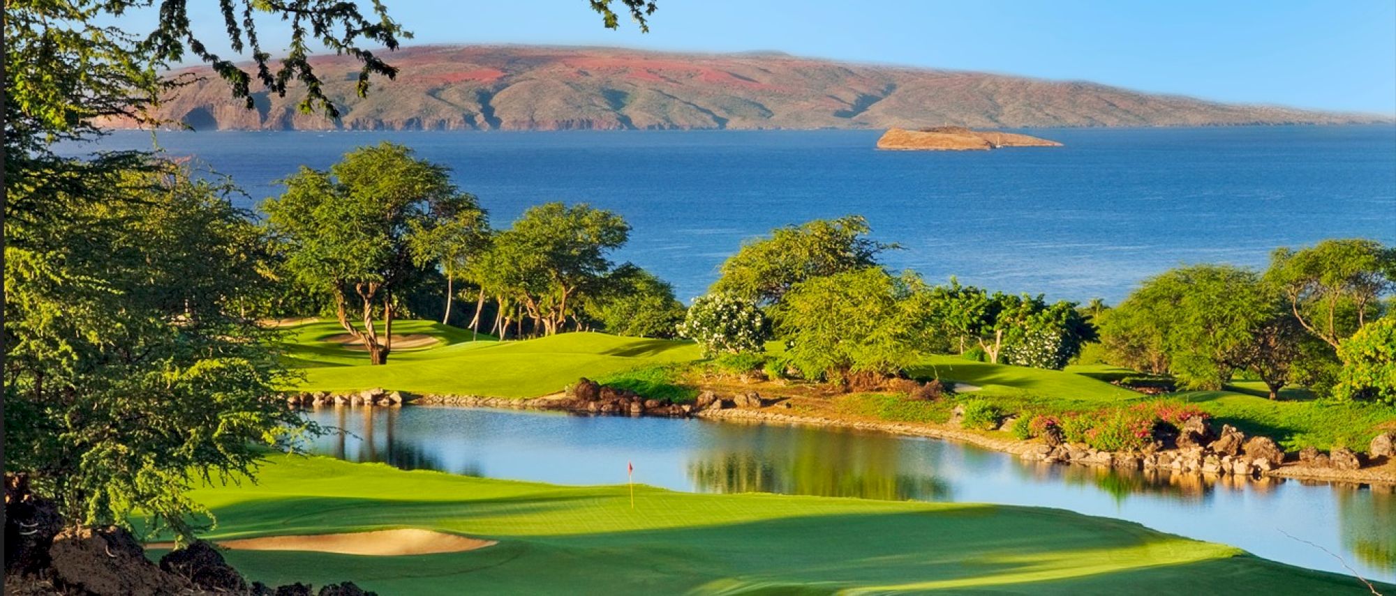 The image shows a beautiful golf course with a lake, lush greenery, and a view of the ocean and distant hills under a clear sky.