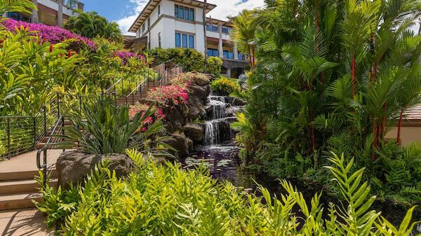 This image shows a picturesque tropical garden with lush greenery, colorful flowers, a cascading waterfall, and a building in the background.