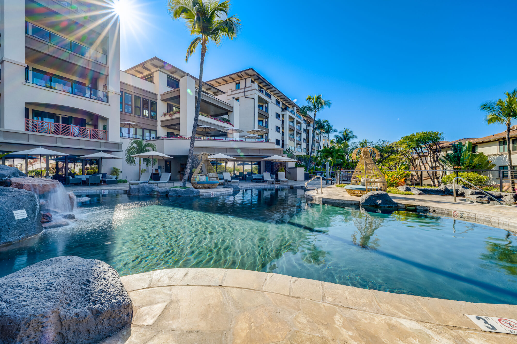 A sunny resort pool area with a large, curved pool, lounge chairs, palm trees, and a multi-story building in the background, bright blue sky, and sunny vibe.
