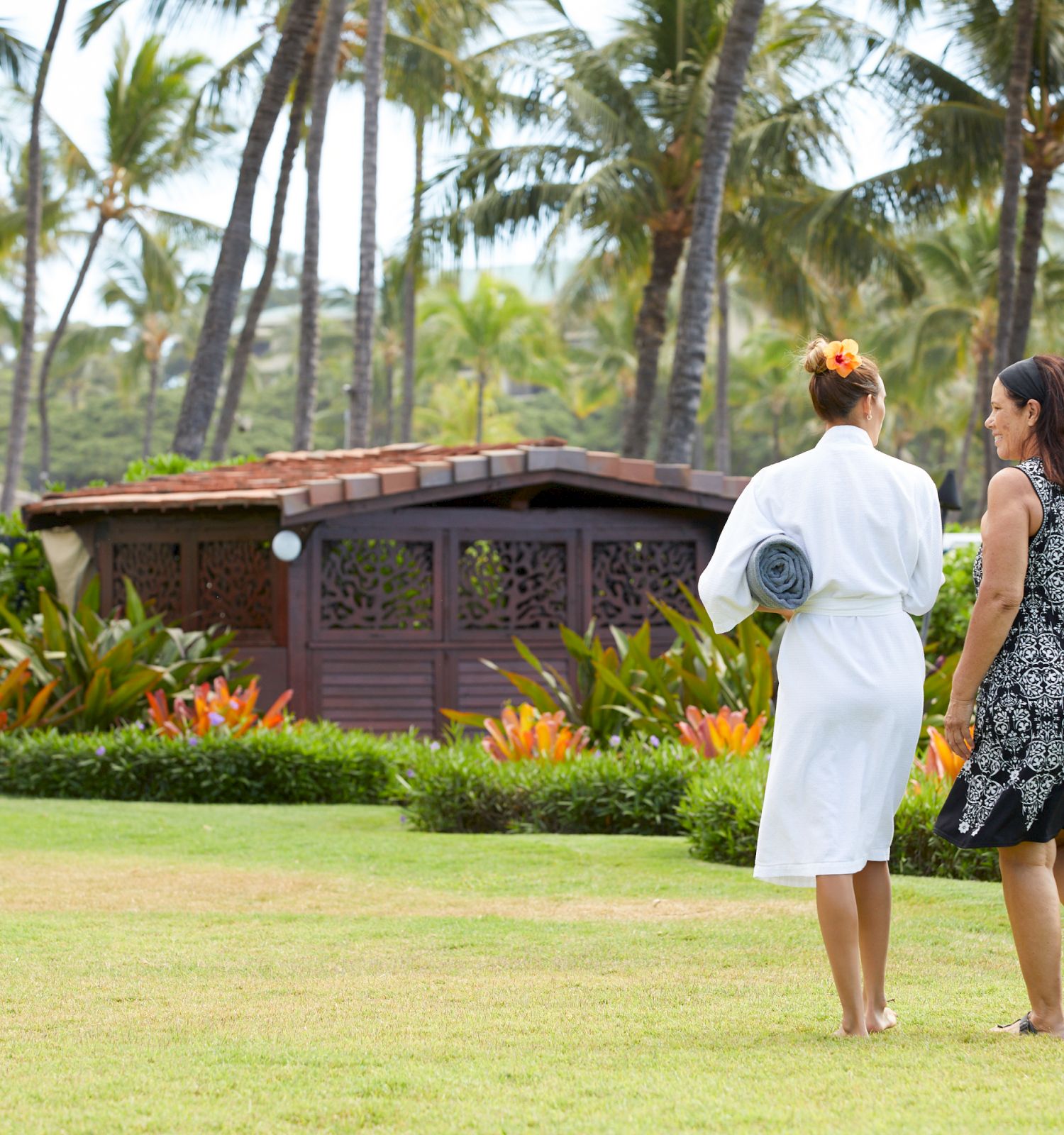 Two people walking on lush green grass towards a wooden building surrounded by tropical plants and palm trees in a serene setting.