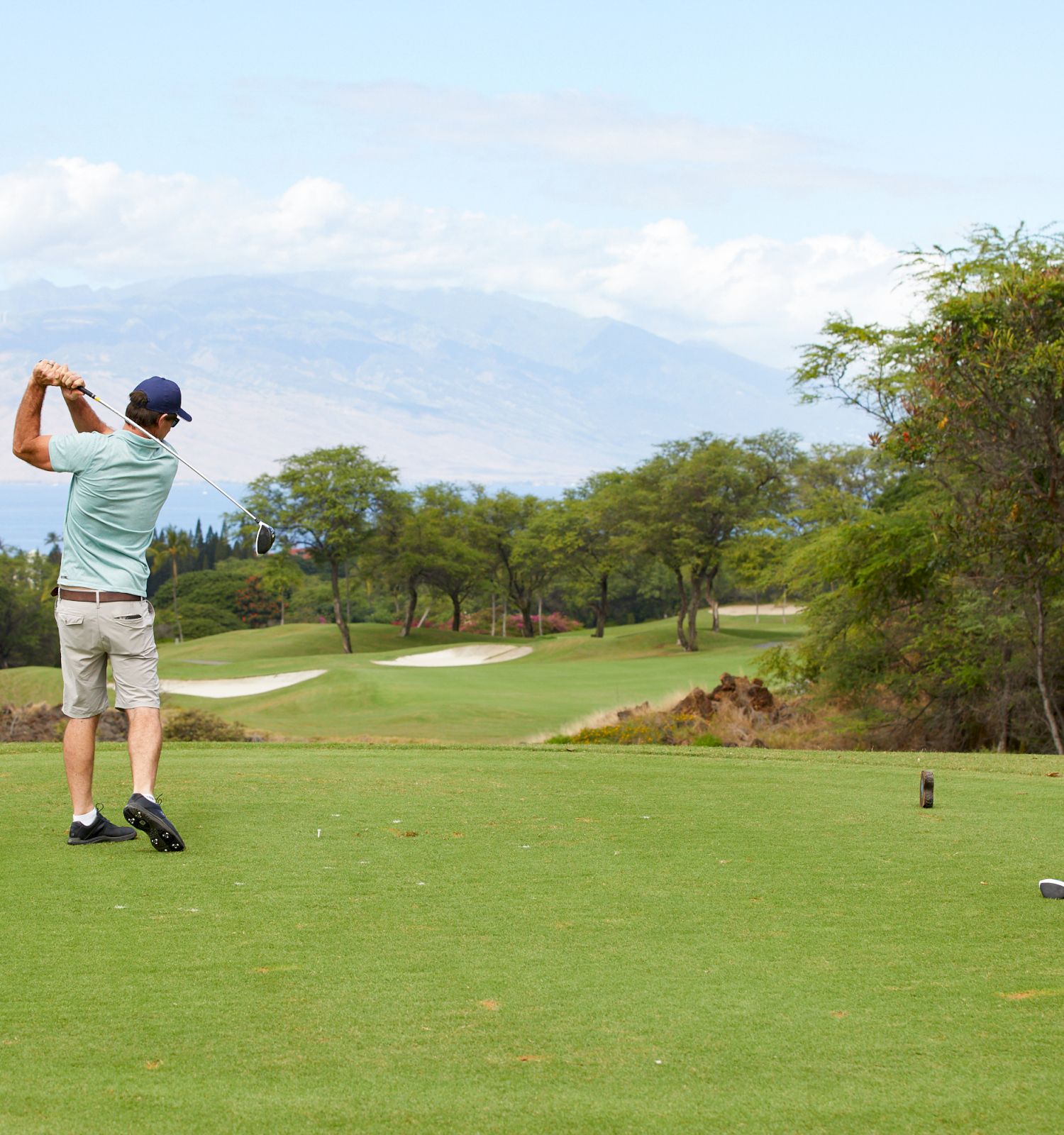 Two people are on a golf course; one is swinging a club while the other watches. The course is lush and scenic with mountains in the background.