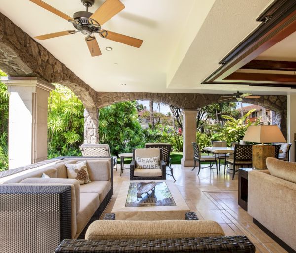 The image shows a stylish outdoor living area with comfortable seating, a ceiling fan, and lush greenery in the background.
