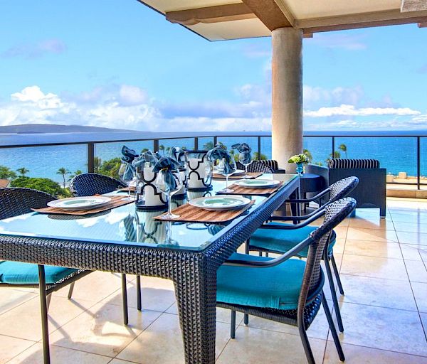 An outdoor deck with a dining table set for a meal, overlooking the ocean. There are lounge chairs on the side and a stunning ocean view.