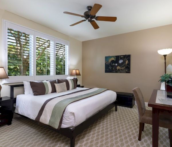 The image shows a cozy bedroom with a large bed, bedside tables, a fan, and a desk, featuring warm colors and natural light.