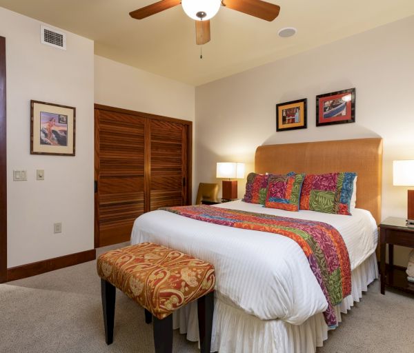 The image shows a cozy bedroom with a large bed, colorful decor, a stool, and a ceiling fan, creating a warm atmosphere.
