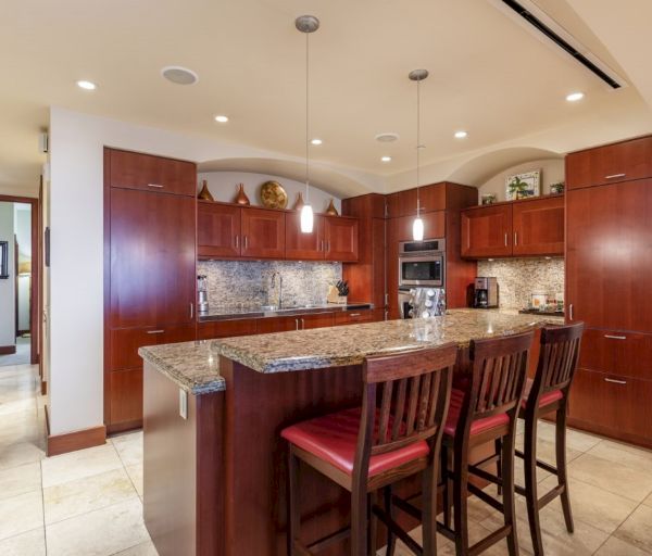 The image shows a modern kitchen featuring wooden cabinetry, granite countertops, and bar seating with red chairs.