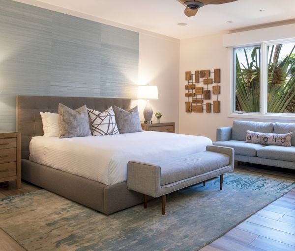 The image shows a modern bedroom featuring a large bed, a cozy sofa, lamps, and a window with greenery outside.