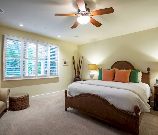 The image shows a cozy bedroom with a bed, colorful pillows, a ceiling fan, and natural light from the windows.