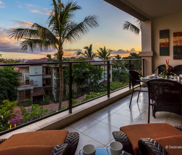 A tropical balcony at sunset with palm trees, outdoor seating, and a view of nearby buildings and lush greenery.