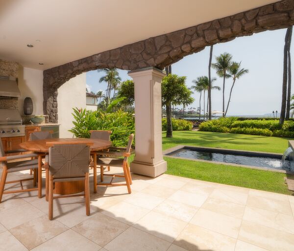 A covered outdoor patio with a dining table and chairs, stone arches, a grassy yard, palm trees, and a glimpse of the ocean in the background.