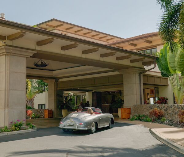 A luxury car pulls into a hotel porte-coch&egrave;re under a covered entrance, tropical landscaping and palm trees lining the driveway.