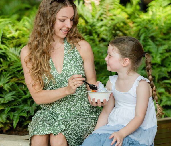 A woman and a young girl sit on a bench outdoors, sharing a bowl of snacks together with smiles.