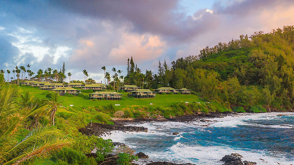 Lush coastal village on a rocky shoreline with green hills, palm trees, and soft waves crashing, colorful sunset clouds overhead.