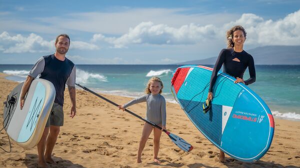 A family at the beach with two adults and a child carrying surfboards and a paddleboard, ready to surf by the ocean.