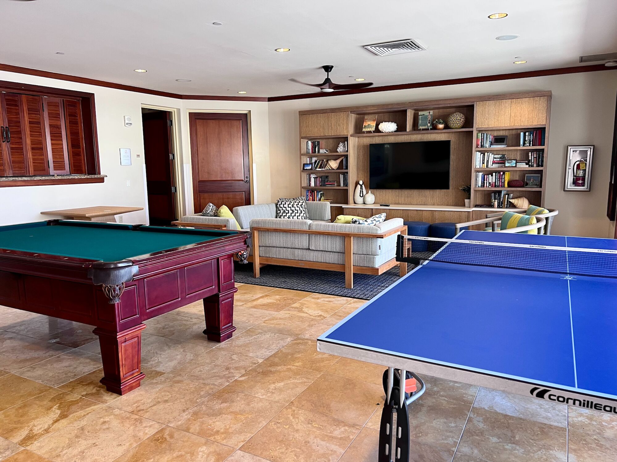 A spacious rec room with a pool table, a blue ping-pong table, beige seating area, built-in shelves, and a large TV console in the background, neat and inviting.