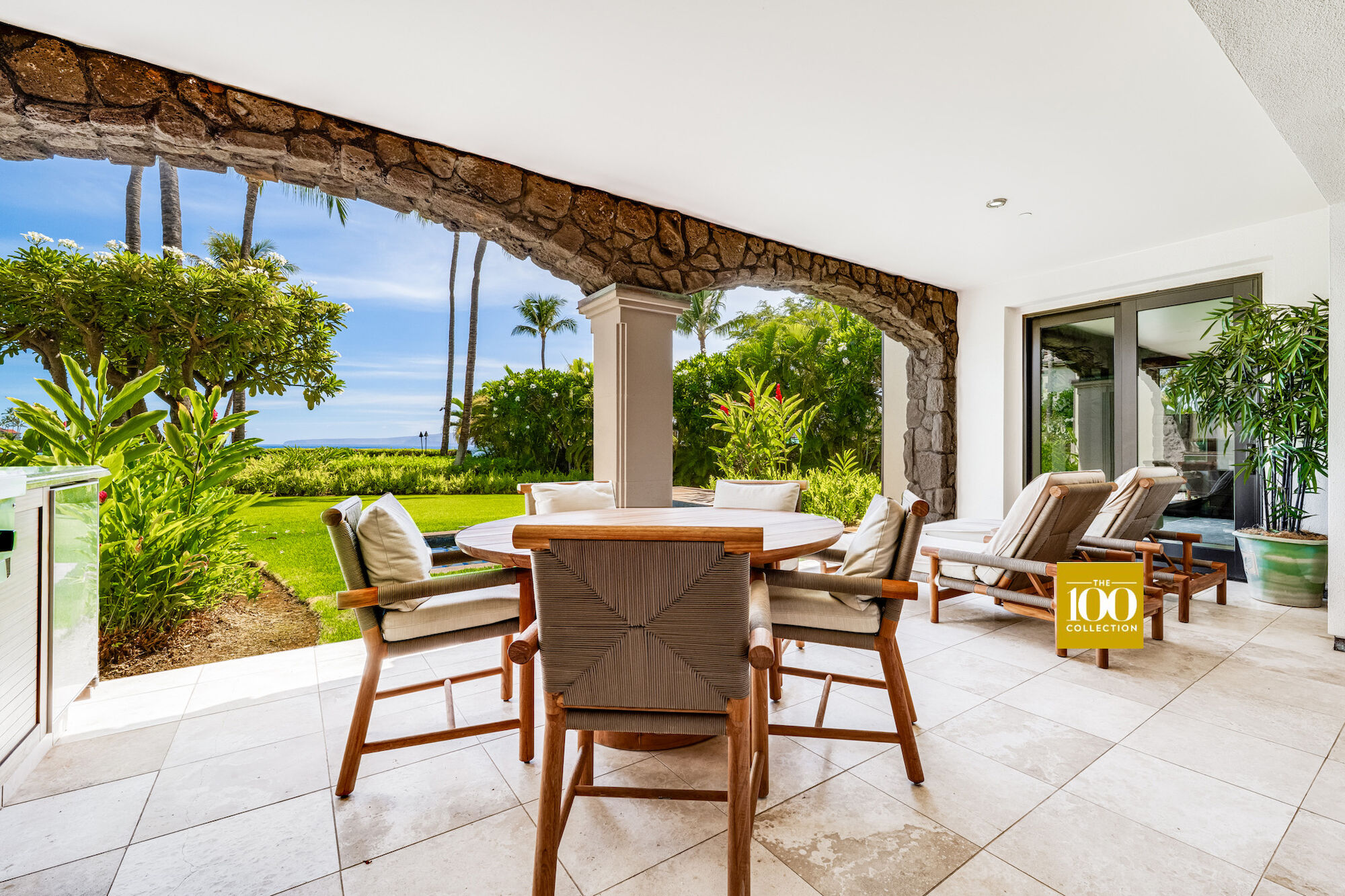 A covered patio with a round dining table and six chairs, lush tropical plants, palm trees, and a sunny lawn beyond the arches.