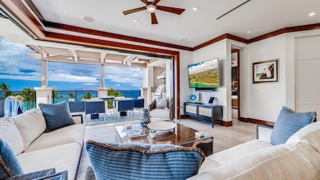 Luxurious open living room with coastal views, white and blue decor, plush seating, and a TV; outdoor patio beyond, sunny sky.