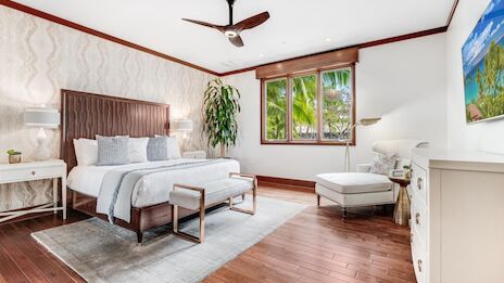 A bright, stylish bedroom with a wooden floor, king bed, nightstands, a bench, plant, window seating, ceiling fan, and minimal d&eacute;cor.