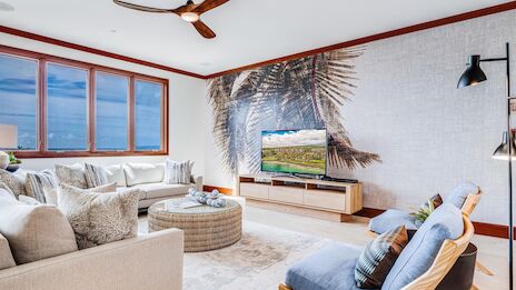 A bright, airy living room with a beachy vibe: light sofa, a round ottoman, wall mural of palm trees, wooden TV stand, large window, and a blue accent chair with a floor lamp, all under a ceiling fan, and a calm, coastal feel.