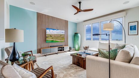 A bright coastal living room with a large TV, teal accent wall, mid-century chairs, a wooden coffee table, and a ceiling fan; sunny windows.