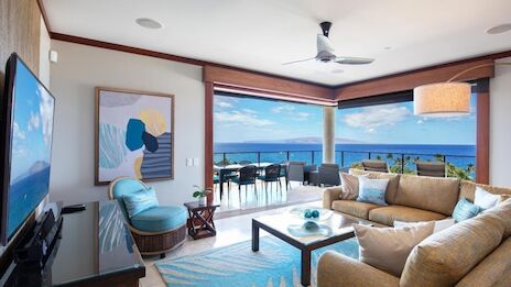 A bright, modern living room with ocean views, comfortable tan sofas, teal accents, a glass coffee table, and a balcony dining area overlooking the sea.