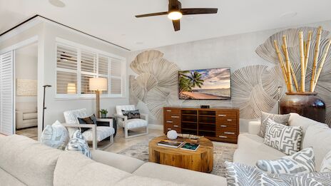 A bright, modern living room with a large beige sectional, tropical decor, a wooden media console, coffee table, and ceiling fan&mdash;cozy yet stylish.