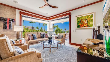 A bright, airy living room with wicker sofas, a glass coffee table, palm view windows, a ceiling fan, art on the walls, and a console with decor, ending with a period.